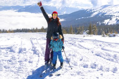 Genç sporcu kadın dağlarda kayak yapan çocukla kayak yaparken eğleniyor. Küçük anne oğul ailesi ekstrem spor aktivitelerinin tadını çıkarıyor.