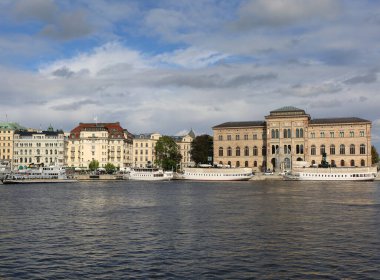 Stockholm, İsveç-18 Eylül 2025: Stromkajen Limanı 'ndaki tarihi binaların önünde demirlemiş beyaz feribot ve gezi tekneleri ve güzel mavi bulutlu gökyüzüyle yürüyen kimliği belirsiz insanlar