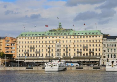 Stockholm, İsveç-18 Eylül 2025: Renkli Bayraklı Grand Otel Binası, Yürüyen İnsanlar, Demirlemiş Tekneler ve Önünde Tur Otobüsleri
