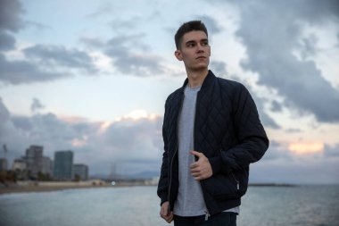 Ceketli genç adam yan bakıyor, şehir ve su arka planda.