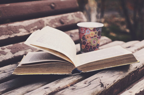 Open book and hot drink on wooden bench in autumn park