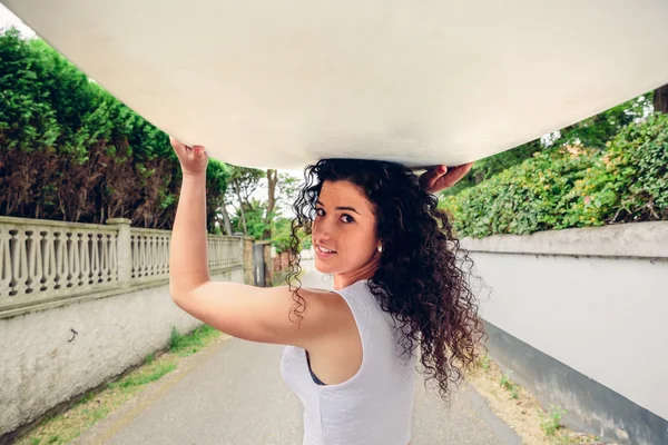 Sonriente chica morena sosteniendo tabla de surf sobre su cabeza — Foto de Stock