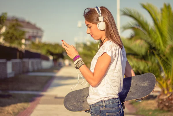 Jong meisje met skateboard en koptelefoon luisteren muziek buitenshuis — Stockfoto