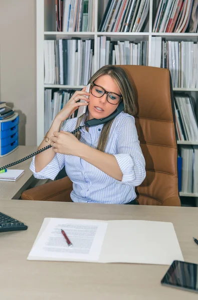 Busy businesswoman talking with cell and fixed line phone in office — Stock Photo, Image