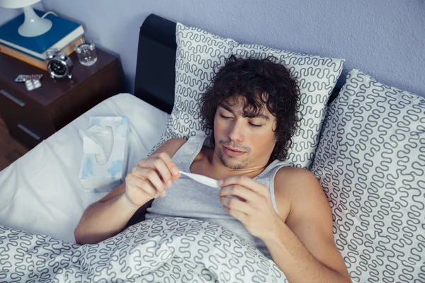 Hombre enfermo buscando temperatura en el termómetro en la cama —  Fotos de Stock