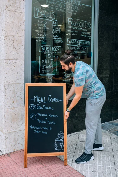 Young waiter placing a blackboard with the daily menu outdoors — Stock Image Young waiter placing a blackboard with the daily menu outdoors Royalty Free Stock Images