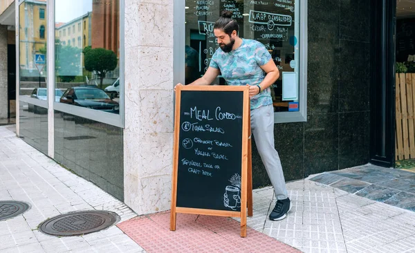 Garçom jovem colocando um quadro negro com o menu diário ao ar livre — Fotografia de Stock