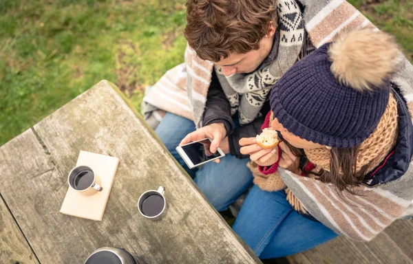 Young couple under blanket looking smartphone and eating muffin outdoors — Stock Photo, Image