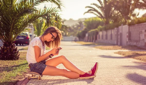 Kaykay ve açık havada yaz üzerinde oturan smartphone ile genç kız — Stok fotoğraf