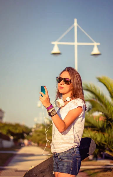 Mladá dívka s skateboard a sluchátka hledět smartphone — Stock fotografie