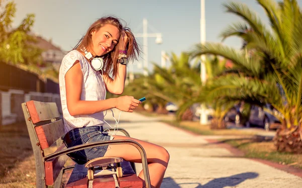 Chica joven con monopatín y auriculares que buscan teléfono inteligente — Foto de Stock