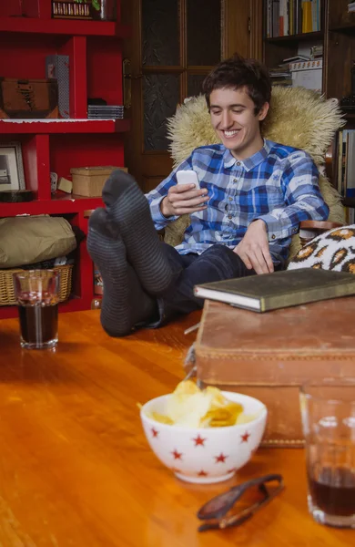 Ung man skrattar och ser smartphone sitter på stol — Stockfoto