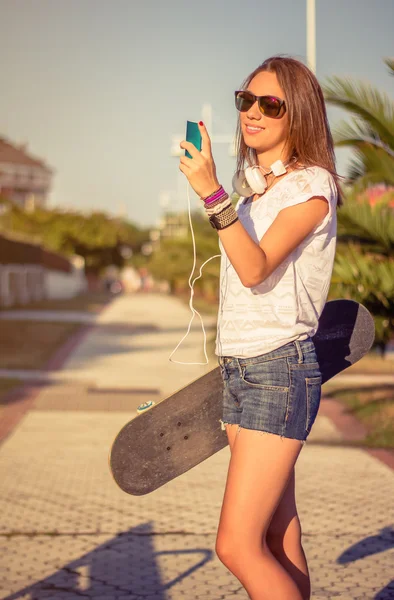 Mladá dívka s skateboard a sluchátka hledět smartphone — Stock fotografie