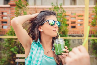 Woman with sunglasses drinking green vegetable smoothie outdoors clipart