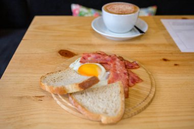 Geleneksel kahvaltıda yumurta, kızarmış domuz pastırması, bir dilim ekmek ve bir restoranda ya da kafede hafif ahşap masada servis edilen sıcak bir kapuçino ile sıcak bir sabah sahnesi yaratılır.