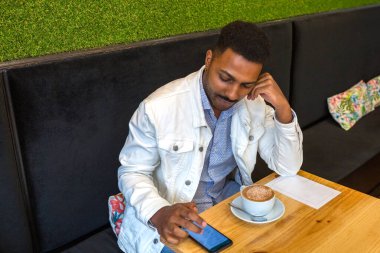 Genç adam modern bir kafede dinleniyor, teknoloji ile uğraşırken cappuccino içiyor ve akıllı telefonunda geziniyor, boş zamanları ve bağlı bir yaşam tarzını temsil ediyor.