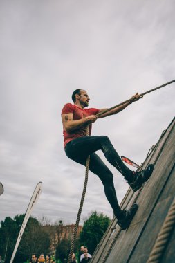 Runner climbing wall with a rope in test of extreme obstacle race