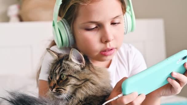 Een tiener met koptelefoon luistert naar muziek, houdt een telefoon in zijn handen en knuffelt een kat op het bed. Het concept van hobby 's, het gebruik van sociale netwerken, moderne gadgets door kinderen. Langzame beweging — Stockvideo