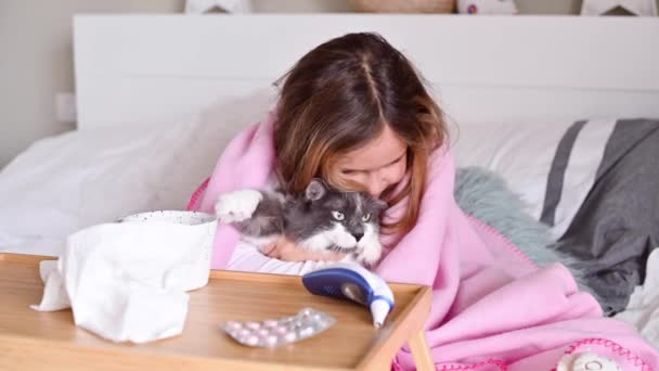 Das kleine Mädchen ist krank. Welpe sitzt auf dem Bett, umarmt und spielt mit der Katze. Auf dem Bett liegt ein kleiner Tisch mit Medikamenten, einem Thermometer und Servietten. — Stockvideo