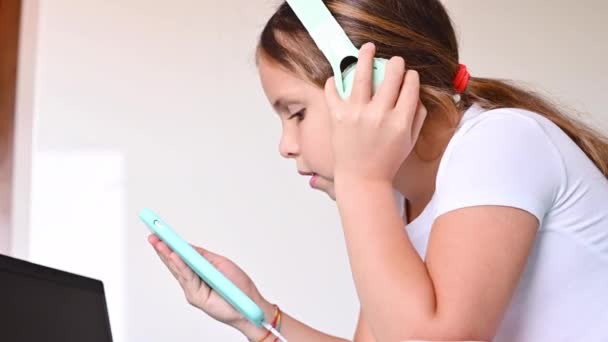 Uma adolescente com fones de ouvido ouvindo música e segurando um telefone em suas mãos, ela está no quarto. O conceito do uso de redes sociais, gadgets modernos por crianças. Espaço de cópia — Vídeo de Stock