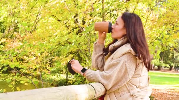 Дівчина з маскою і кавою в екологічній упаковці відпочиває в осінньому парку. Молода жінка з кавою на відкритому повітрі . — стокове відео
