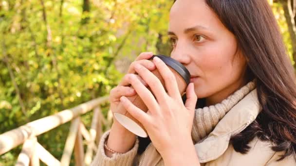 A menina bebe café no parque de outono. Uma jovem mulher com um café takeaway em um pacote ecológico caminha no espaço aberto. Face e caneca close-up. — Vídeo de Stock