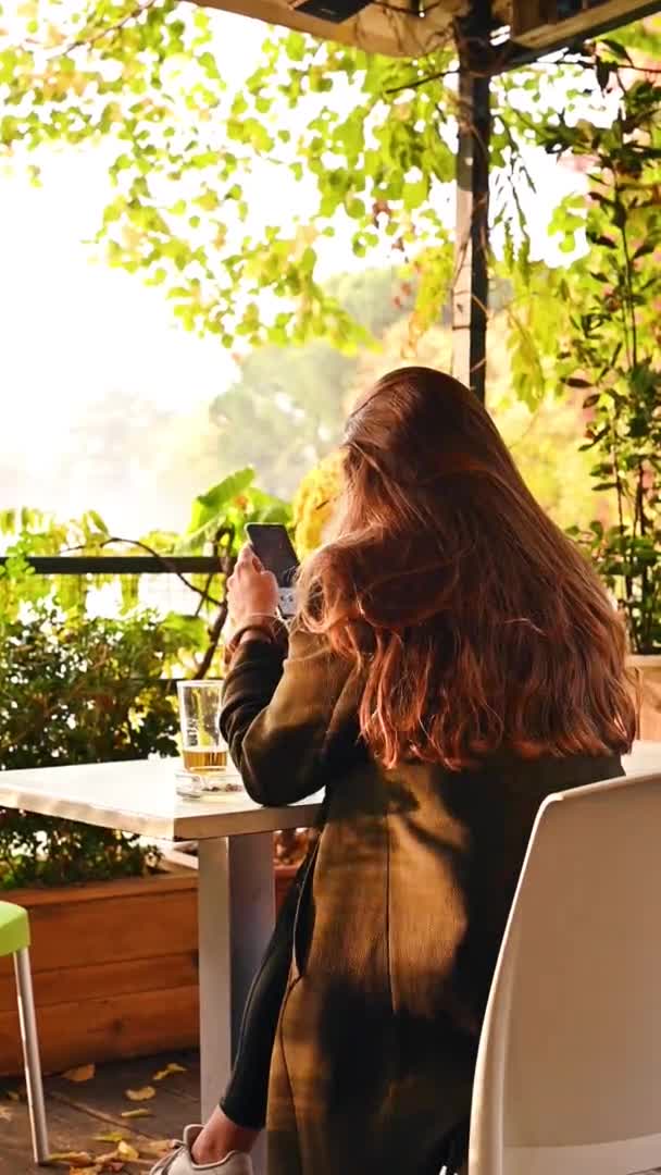 O fată cu un telefon într-o cafenea în aer liber din parc stă la o masă. O femeie tânără cu un martphone în mâini se uită online. Video vertical . — Videoclip de stoc