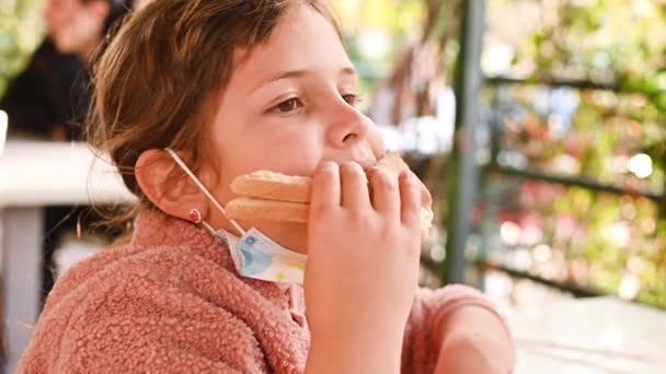 Een klein meisje met een masker eet toast met ham en kaas in een café op straat. Childs gezicht met een sandwich close-up. — Stockvideo