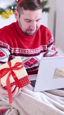 Kırmızı Noel kazağı giyen bir adam evden sürpriz internet siparişi veriyor. İnternetten noel hediyesi almak - online alışveriş konsepti. Yeni yıl tatili konsepti. Dikey video. 4k görüntü