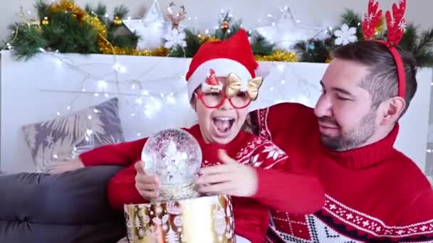 Una bambina con un cappello da Babbo Natale ha preso una palla di vetro da papa '. Famiglia felice a Natale si scambia regali a casa. Bambini e genitori in vacanza. — Video Stock