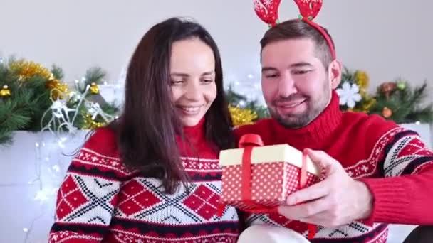 Casal feliz no amor em camisolas vermelhas de Natal em uma casa decorada com guirlandas brilhantes. Um homem dá um presente à sua amada mulher. Feliz Ano Novo conceito. — Vídeo de Stock