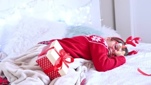 Petite fille dans un pull de Noël avec des cadeaux dans une chambre décorée de guirlandes. Célébration de la nouvelle année chez nous en 2021. Enfant heureux à la maison. — Video