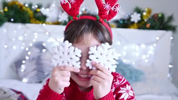 Une petite fille aux émotions joyeuses dans un pull de Noël joue avec les flocons de neige du Nouvel An. Joyeux enfant dans une salle lumineuse festive — Video