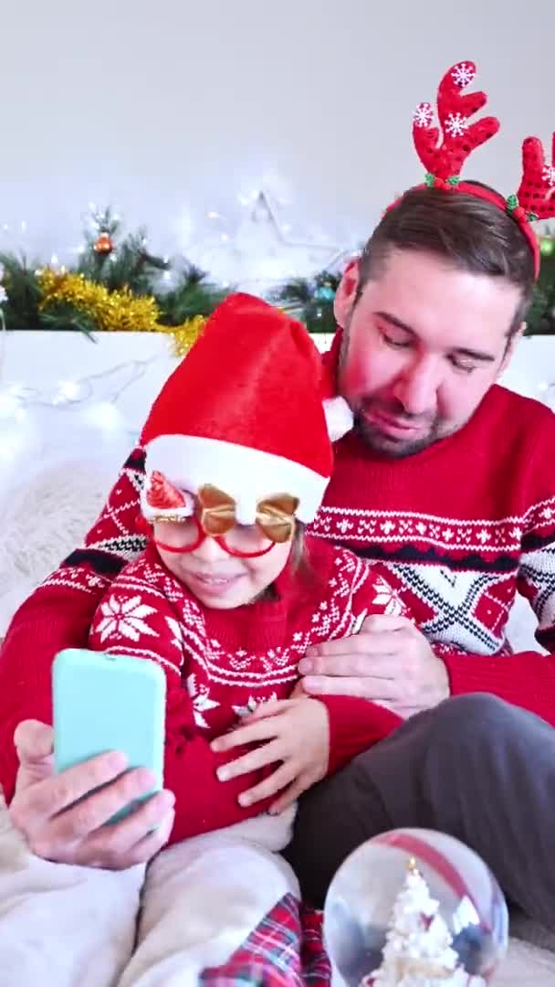 크리스마스 모자를 쓴 아빠와 딸은 메리 크리스마스와 새해를 기념하는 비디오 전화를 하고 있다. 온라인 크리스마스 (Online holiday of Christmas) 2021 년 크리스마스. 의사소통을 위한 스마트폰을 가진 행복 한 가족. 4k 필름 — 비디오