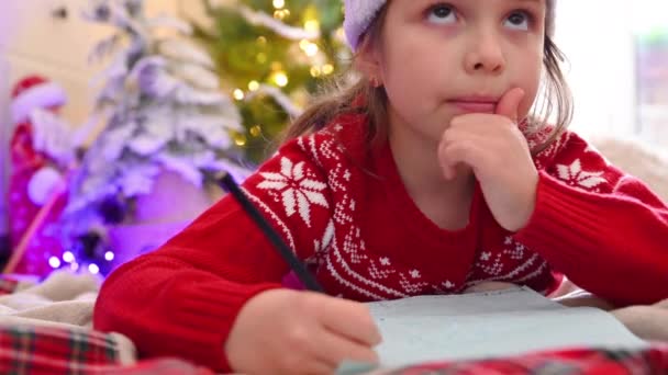 สาวน้อยน่ารักเขียนจดหมายถึงซานตาคลอส เด็กในหมวกและเสื้อสเวตเตอร์สีแดงที่บ้านใกล้ต้นคริสต์มาส . — วีดีโอสต็อก