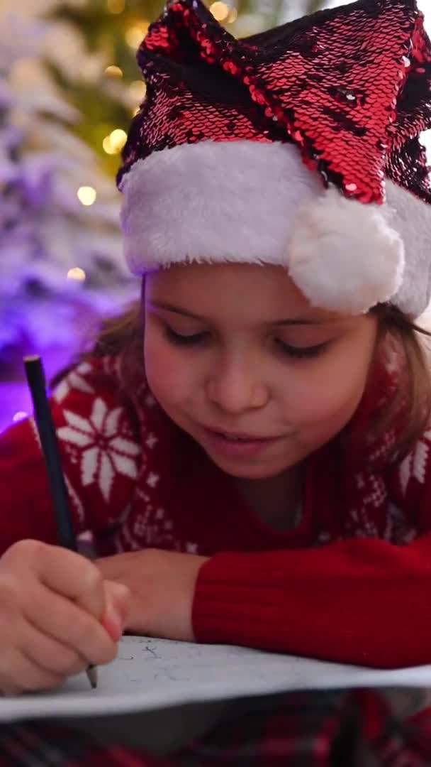 Petite fille écrit une lettre au Père Noël dans la douce maison près du sapin de Noël. L'enfant rêve de cadeaux pour les vacances du Nouvel An et des bougies brûlent sur la table — Video