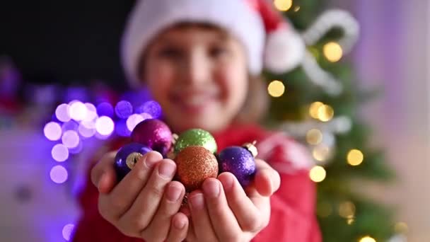 Ein kleines Mädchen schmückt einen Weihnachtsbaum und hat Spaß mit Weihnachtskugeln in der Hand. Glückliches Kind im weihnachtlich geschmückten Haus. — Stockvideo