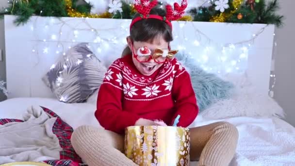 A boldog gyerek otthon ajándékokat nyit. Az új évet otthon ünnepeljük 2021-ben. Egy kislány karácsonyi pulóverben, ajándékokkal egy koszorúval díszített szobában. Kiváló minőségű 4k felvételek — Stock videók