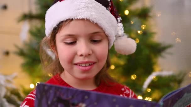 Een klein meisje met een kerstmuts leest een boek bij een kerstboom met bloemenslingers. — Stockvideo