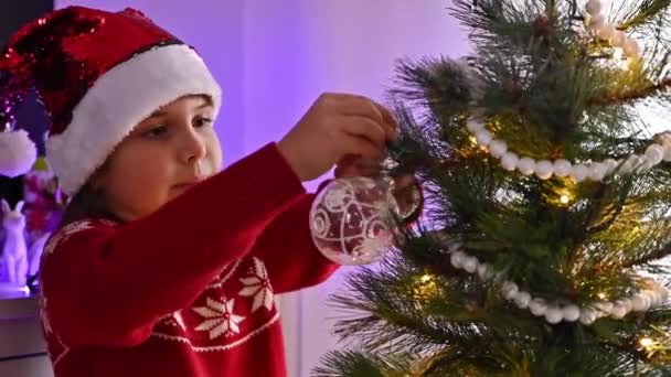 Une petite fille dans un chapeau de Père Noël décore un arbre de Noël dans une maison confortable. Préparation de la célébration de Noël et du Nouvel An. — Video