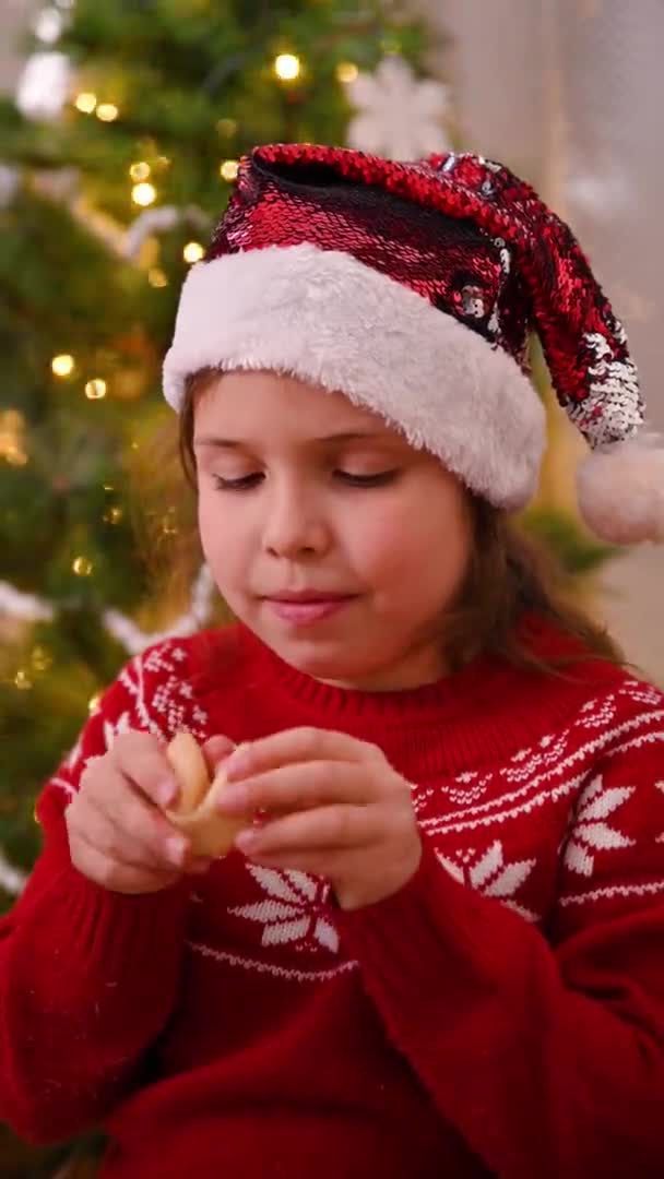 O fetiță cu o pălărie de santaclaus și un pulover roșu lângă pomul de Crăciun deschide o prăjitură cu avere. Tradiția țărilor estice . — Videoclip de stoc