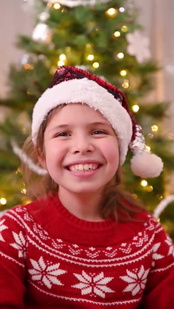 Klein meisje in kerstmuts, rode trui lacht en heeft plezier in een kamer ingericht voor Kerstmis. Verticale video voor smartphones en social media. — Stockvideo