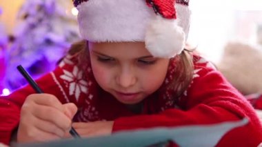 Kırmızı kazaklı küçük bir kız Noel için süslenmiş tatlı bir evde Noel Baba 'ya mektup yazıyor..