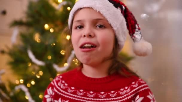 Niña comiendo dulces en Navidad en una hermosa habitación decorada cerca del árbol de Navidad. Lindo bebé en sombrero de santa y suéter rojo en casa en vacaciones. — Vídeos de Stock