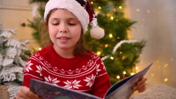 Mała dziewczynka w kapeluszu Mikołaja czyta książkę niedaleko choinki z girlandami. — Wideo stockowe