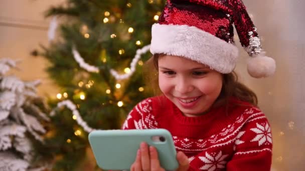 Klein meisje in een rode trui met een telefoon bij de kerstboom in een lief huis. Een kind met een kerstmuts communiceert en heeft plezier online met een smartphone. — Stockvideo