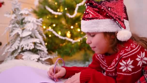 Κοριτσάκι με κόκκινο πουλόβερ που γράφει γράμμα στον Άγιο Βασίλη σε ένα σπιτάκι στολισμένο για τα Χριστούγεννα.. — Αρχείο Βίντεο
