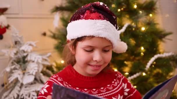 Malá holka v Santově klobouku čte knížku u vánočního stromku s věnci. — Stock video