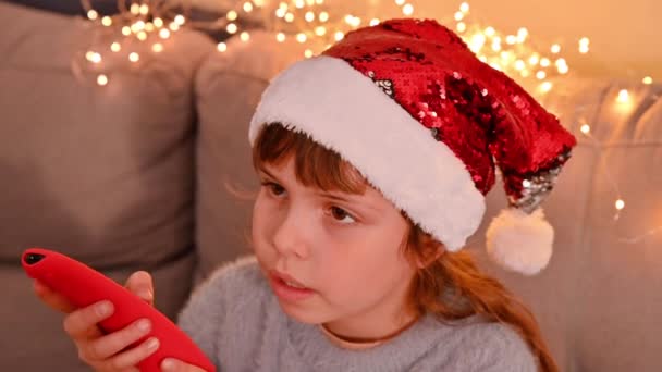 Mała dziewczynka z pilotem ogląda telewizję w pokoju ozdobionym girlandami. Szczęśliwe dziecko w domu na Boże Narodzenie. — Wideo stockowe