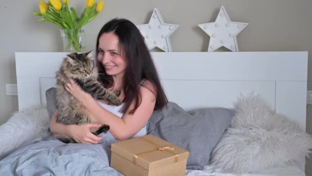 Jonge vrouw in bed in een lichte slaapkamer, knuffelt een schattige kat en een geschenk op het bed. Verrassingsdoos met strik voor moeder of vrouw op 8 maart. — Stockvideo